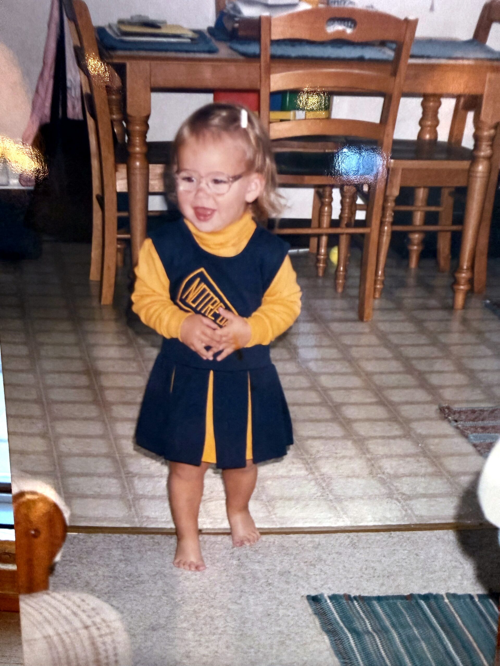 Young child with glasses wearing a navy and yellow outfit, standing indoors on a tiled floor near wooden dining chairs.
