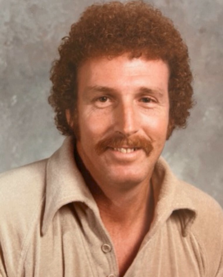 Portrait of a man with curly brown hair and a mustache, wearing a beige collared shirt, smiling at the camera.