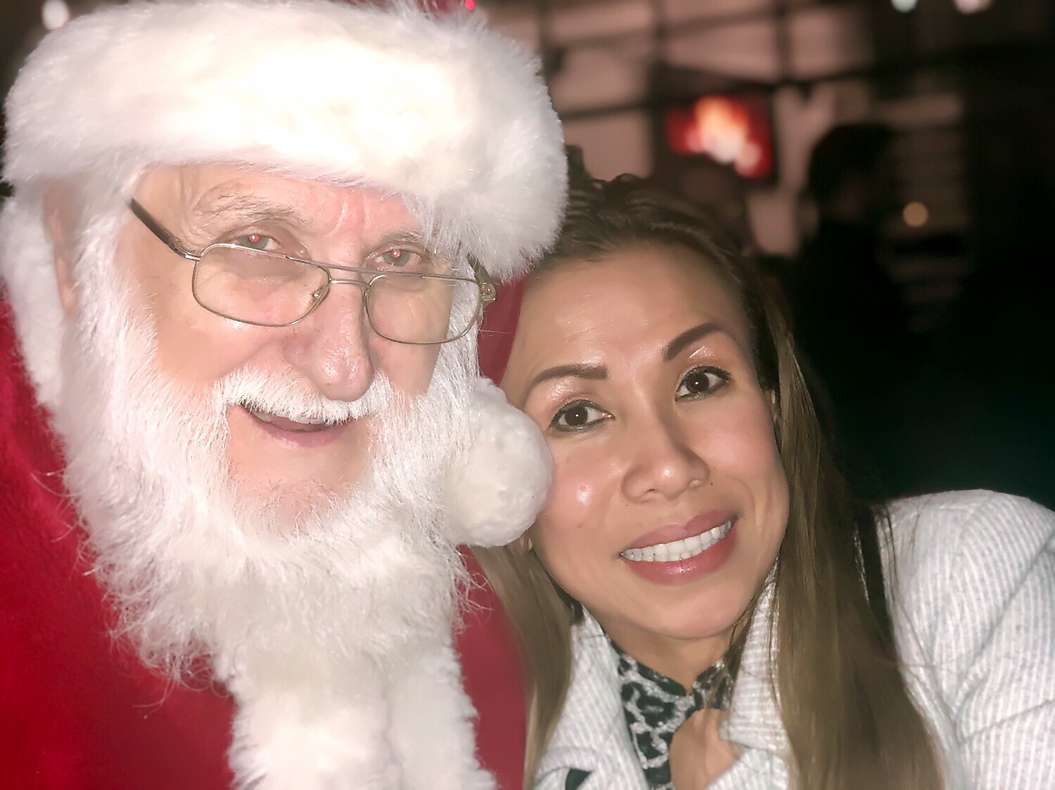 Smiling woman takes a selfie with a man in a Santa Claus costume wearing glasses and a white beard.