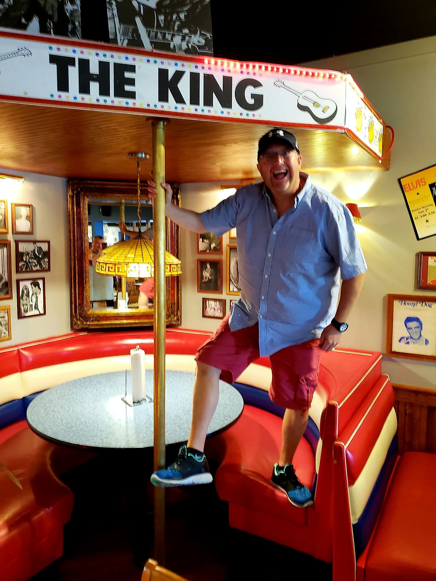 Smiling man in a blue shirt and cap standing on a carnival-style circular ride inside a retro diner.