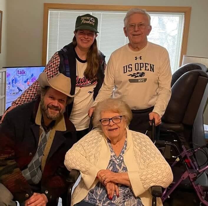 A cheerful group of four posing together indoors: a seated elderly woman in a wheelchair in front, with three adults standing behind her.