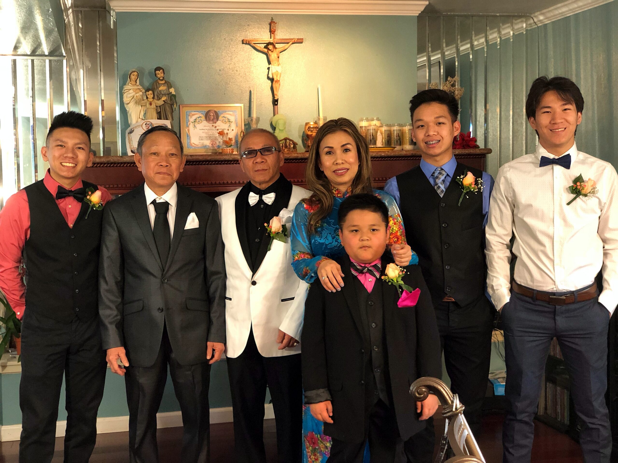 Group of seven people in formal attire posing indoors, with a religious shrine and crucifix behind them.