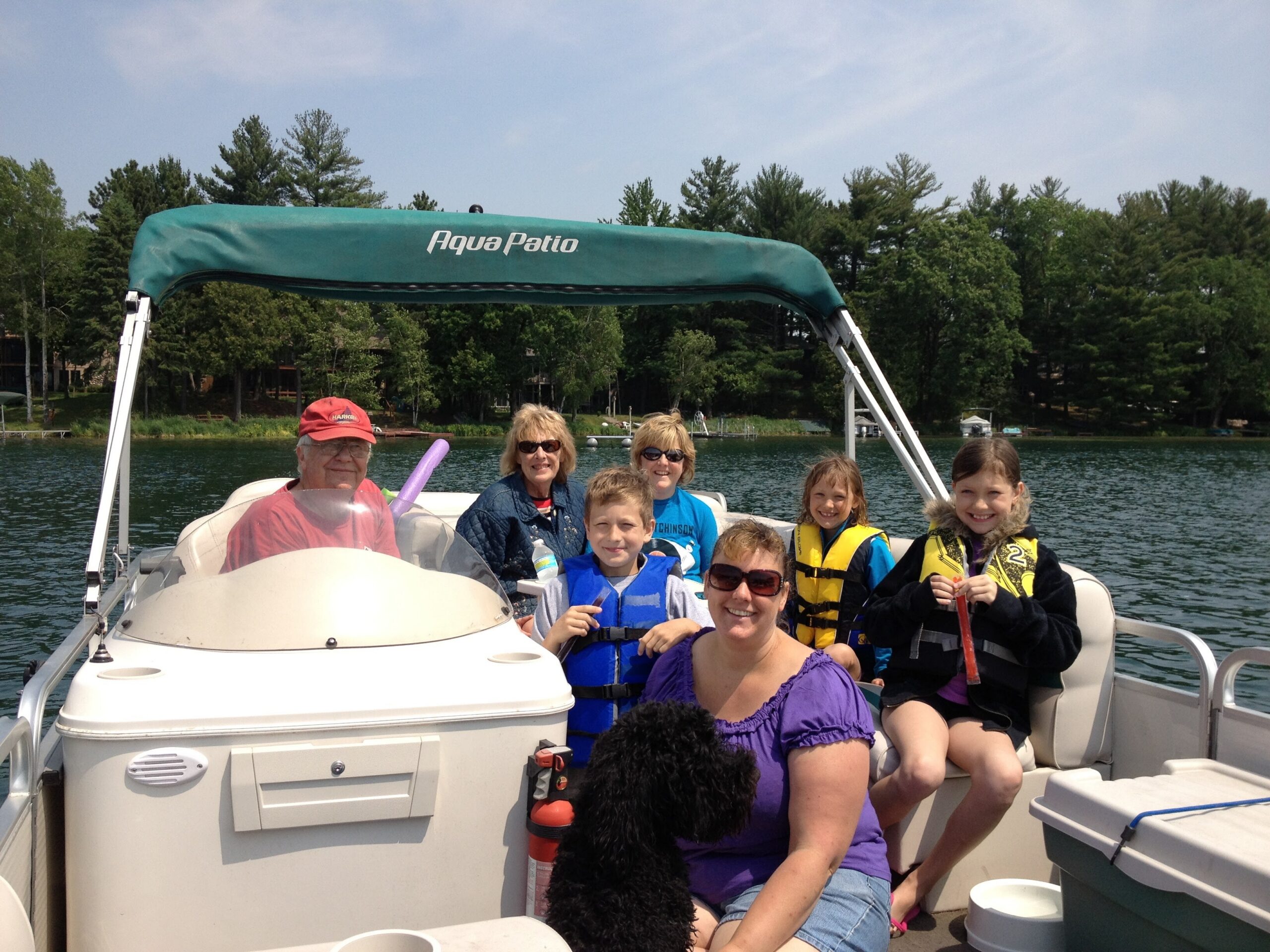 People wearing life jackets sit on a pontoon boat on a lake, smiling, with trees and a blue sky in the background.