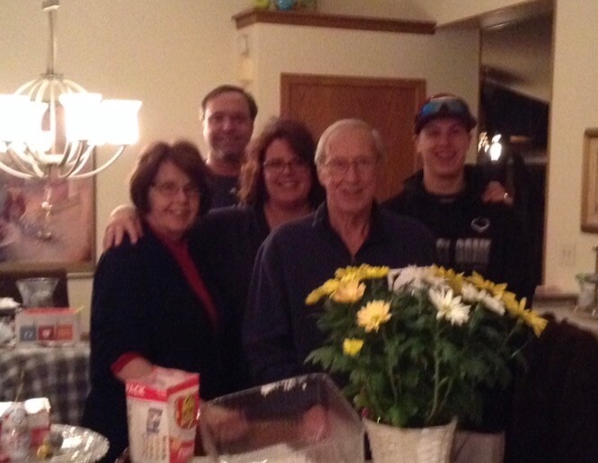 A group of five adults posing together in a home kitchen, smiling around a man with a yellow flower bouquet in front of them.