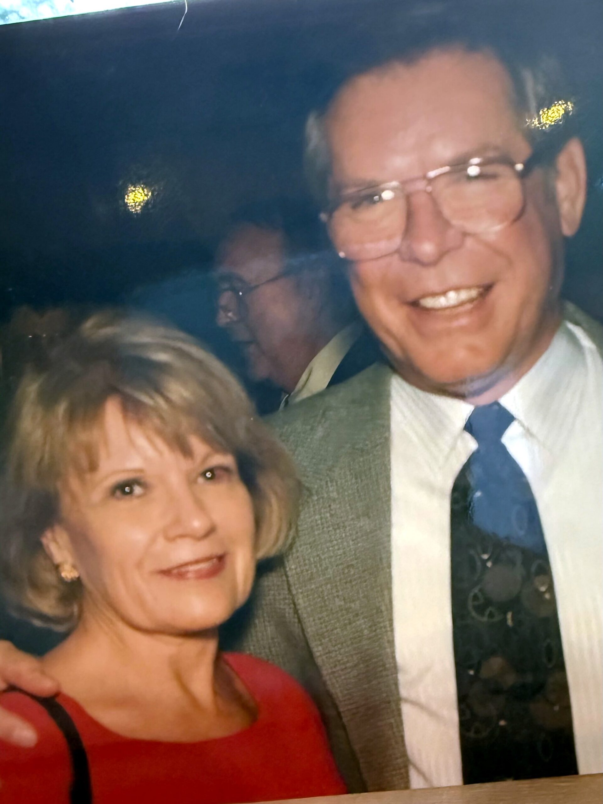 Smiling older woman in a red outfit in the foreground with a man in a suit and glasses beside her.