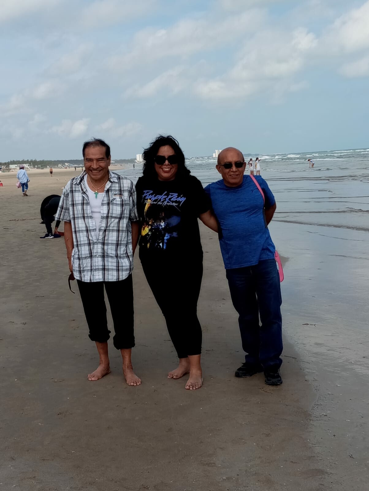 Three adults standing arm-in-arm on a sandy beach with the ocean behind them, smiling at the camera.