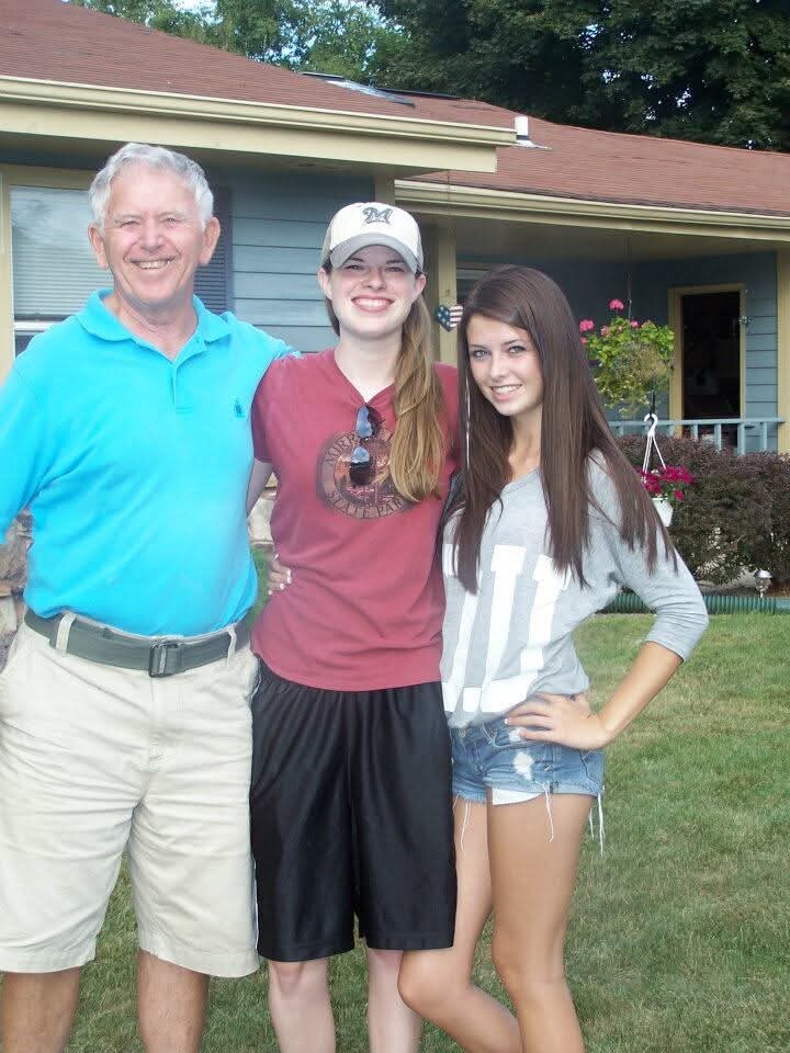 Three people smiling in a front yard in front of a house: an older man in a blue polo, a teen girl in a maroon shirt and cap, and another teen girl in a gray top and denim shorts.