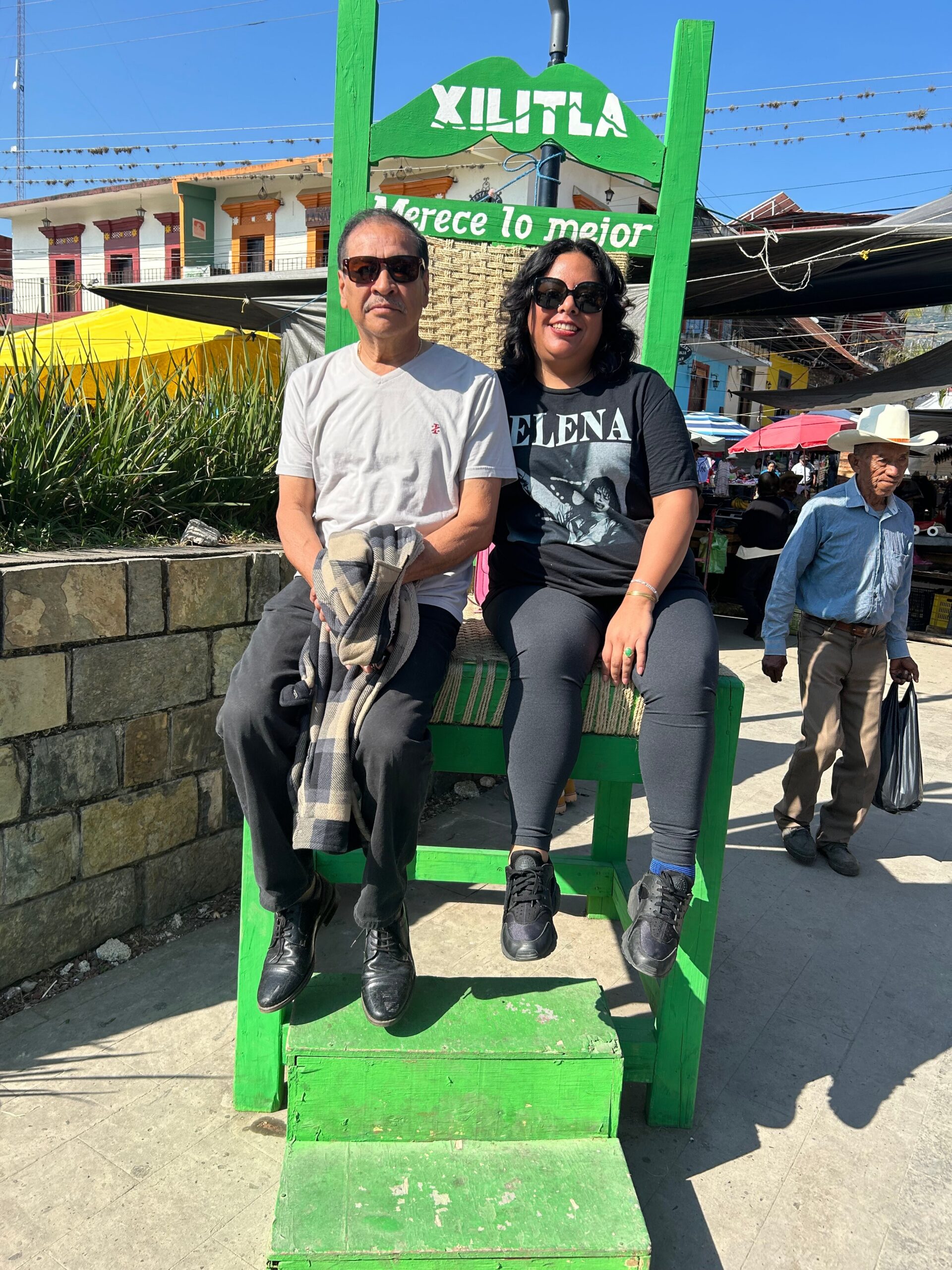 A smiling couple wearing sunglasses sits on a bright green wooden chair in a sunny market square with colorful buildings in the background.