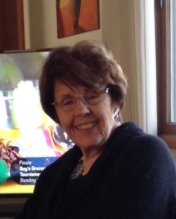 Smiling elderly woman with short brown hair and glasses, wearing a dark sweater indoors by a window with a TV in the background.