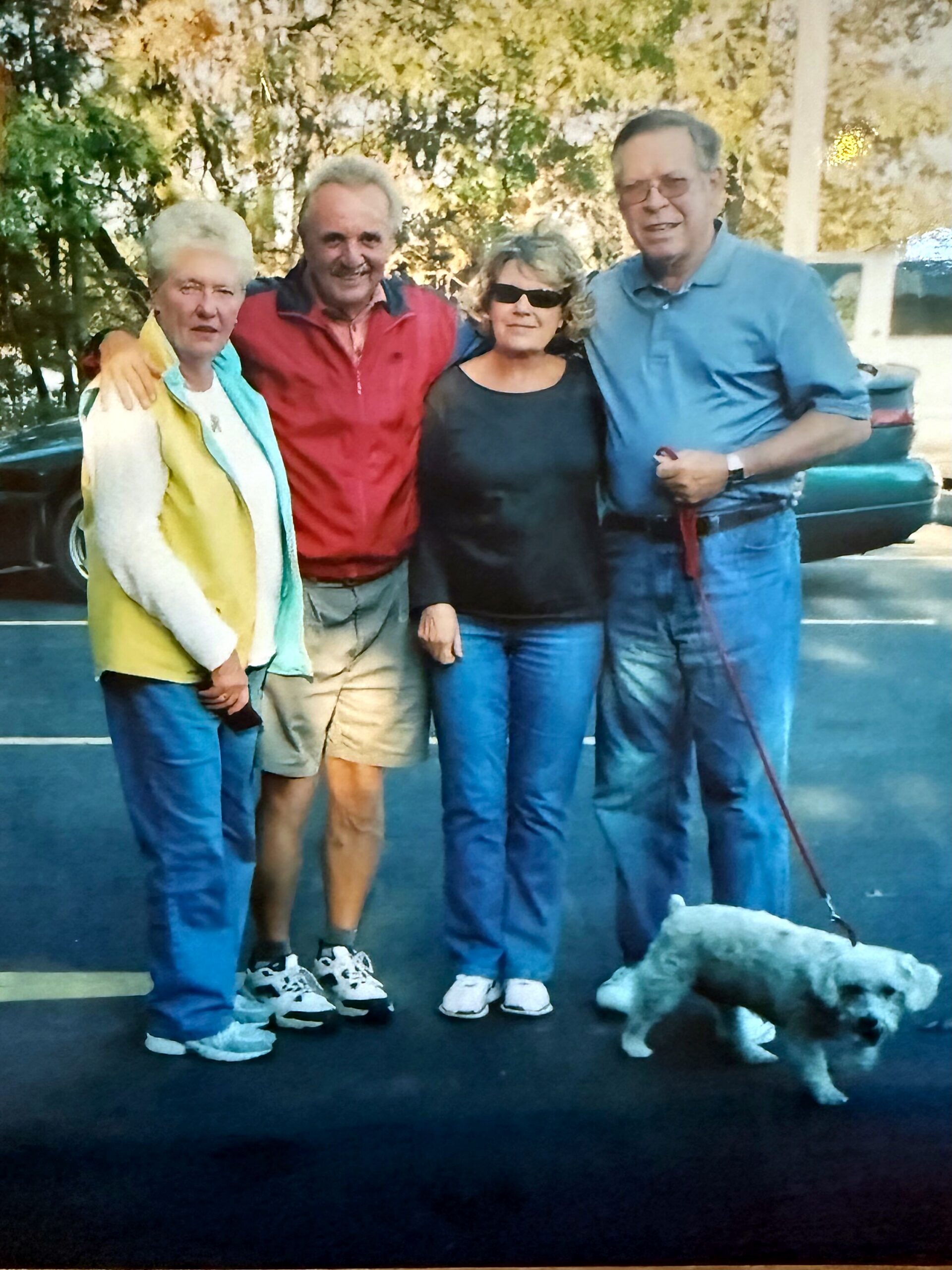 Four adults posing in a parking lot outdoors, smiling, with a small white dog on a leash near the bottom right.