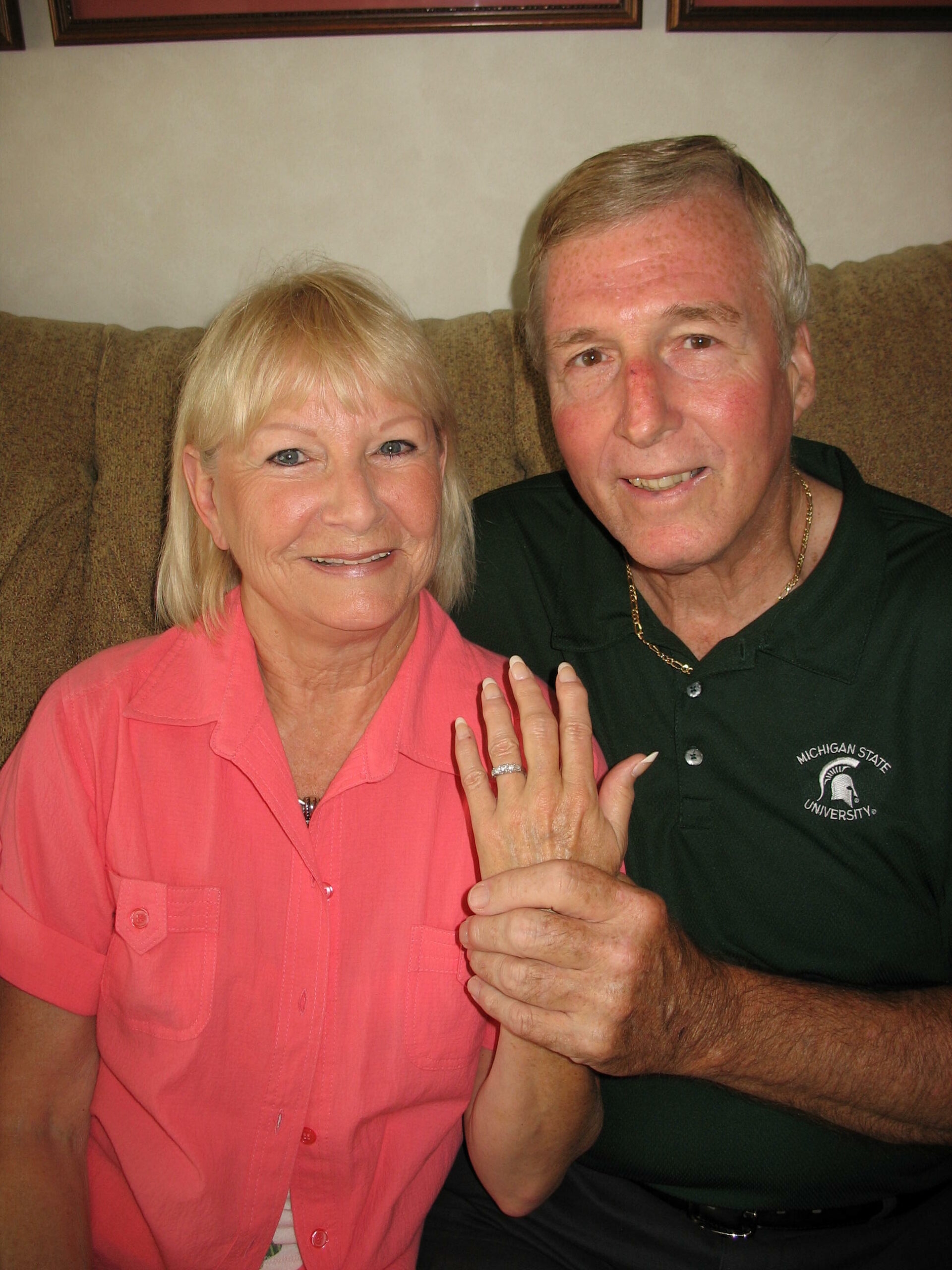 Smiling couple on a couch; woman in pink shirt shows her left-hand engagement ring to the camera.