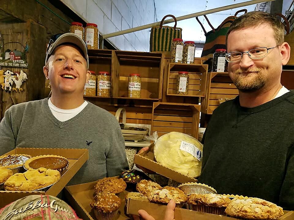 Two men in a bakery hold boxes of assorted pastries and muffins, smiling at the camera.