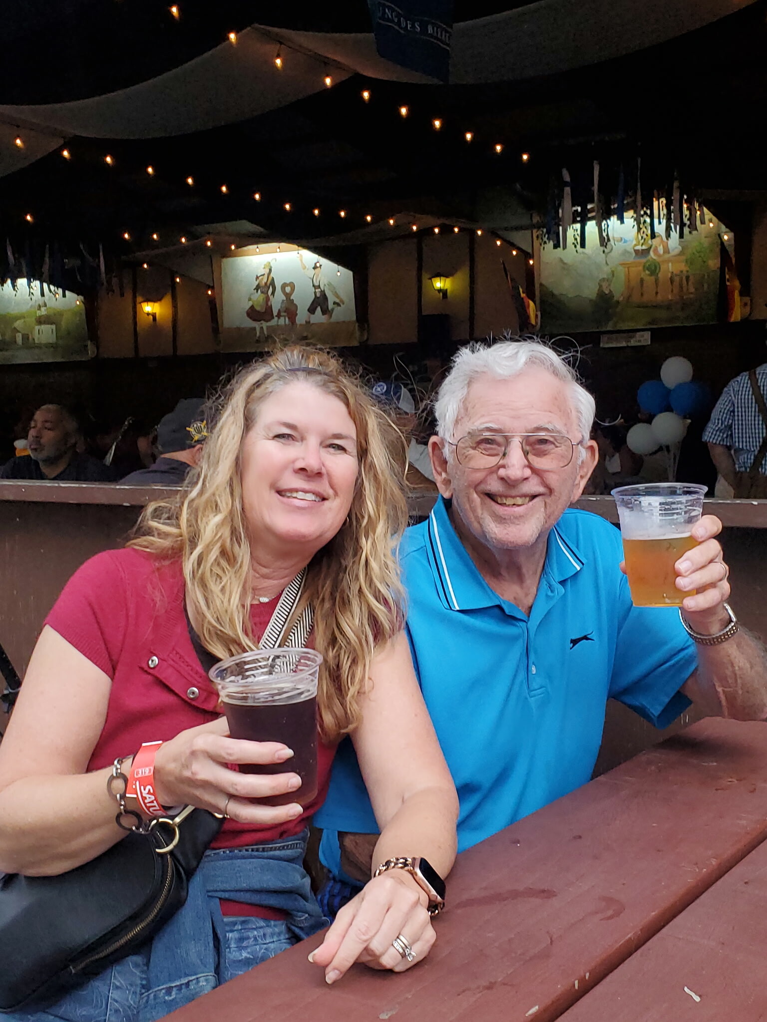 Smiling woman and man sit at a wooden picnic table at night, each holding a plastic cup of beer under string lights in a festival setting.