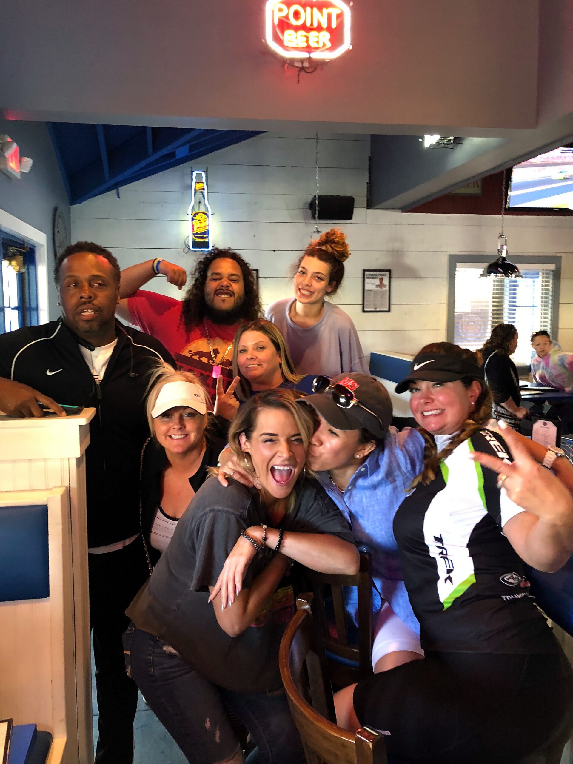 A cheerful group of friends posing closely for a photo inside a casual restaurant with a neon sign overhead, smiling and embracing.