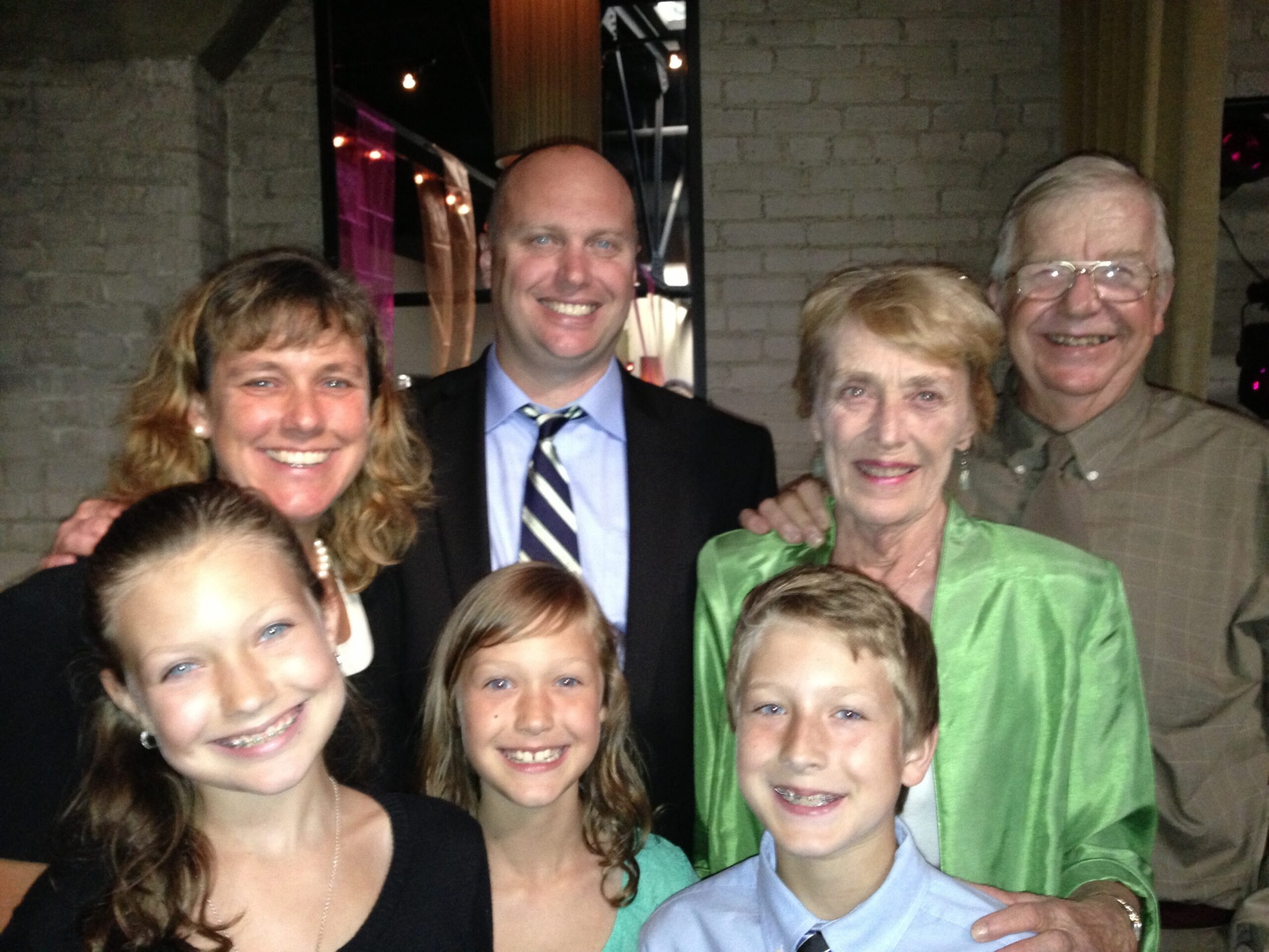 Six people smiling and posing together indoors in front of a brick wall.