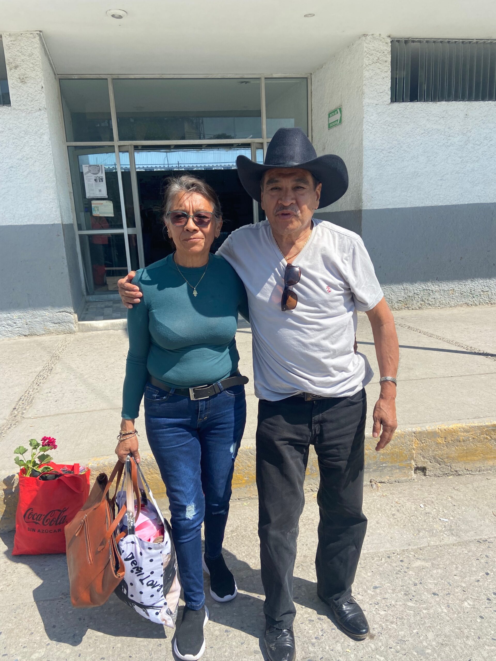 Two adults standing side by side outside a light-gray building, arms around each other, with shopping bags on the ground in front of them.