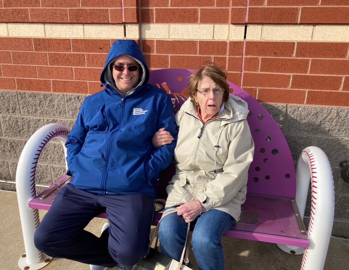 Two seniors sit side by side on a purple bench shaped like a baseball, smiling at the camera. They wear jackets and sunglasses outdoors.