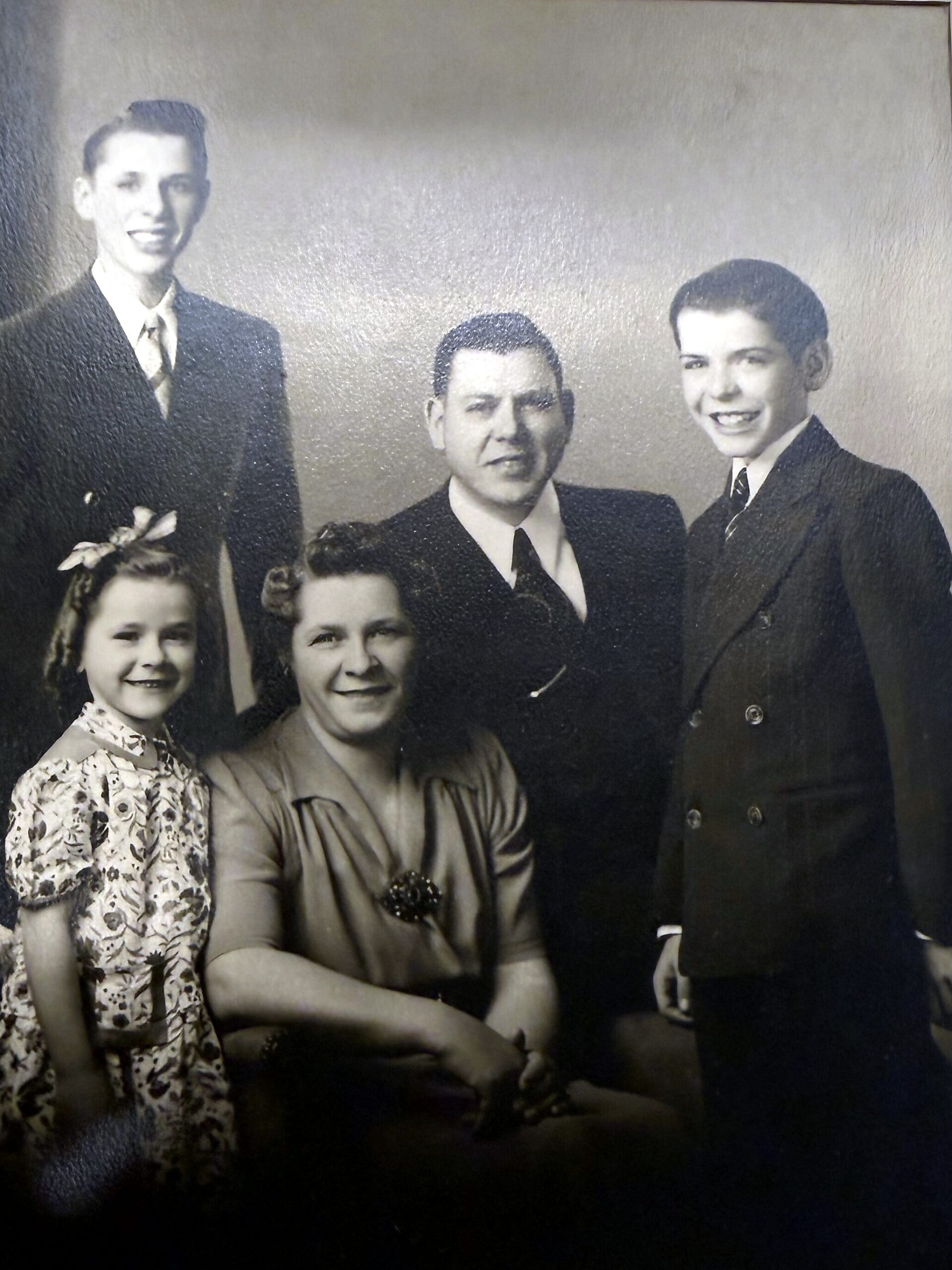 Vintage black-and-white family portrait: three men in suits standing behind two smiling girls seated in front, all dressed formally.