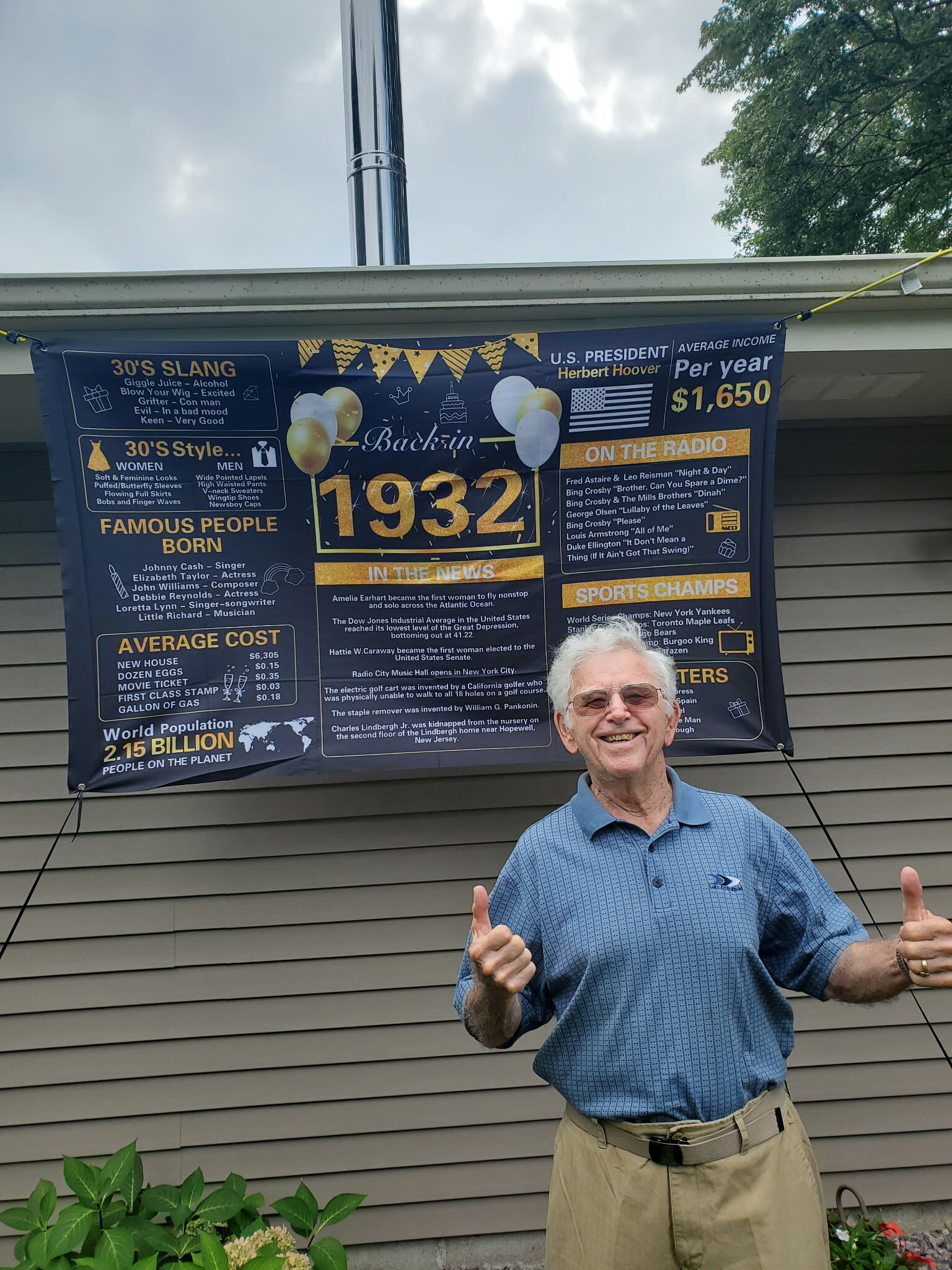 Smiling man in a blue check shirt giving two thumbs up in front of a large blue infographic banner on a house wall.