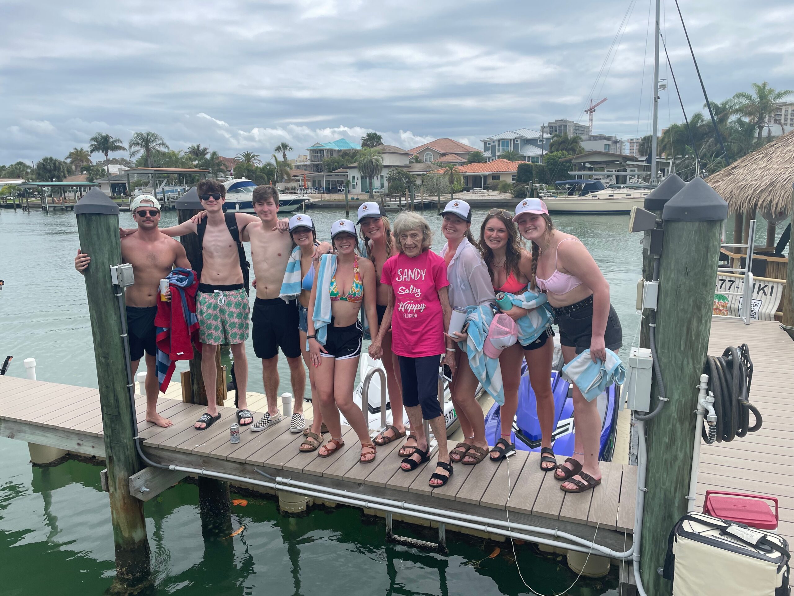 Group of people in swimsuits and hats posing on a marina dock with boats and houses in the background, smiling at the camera.