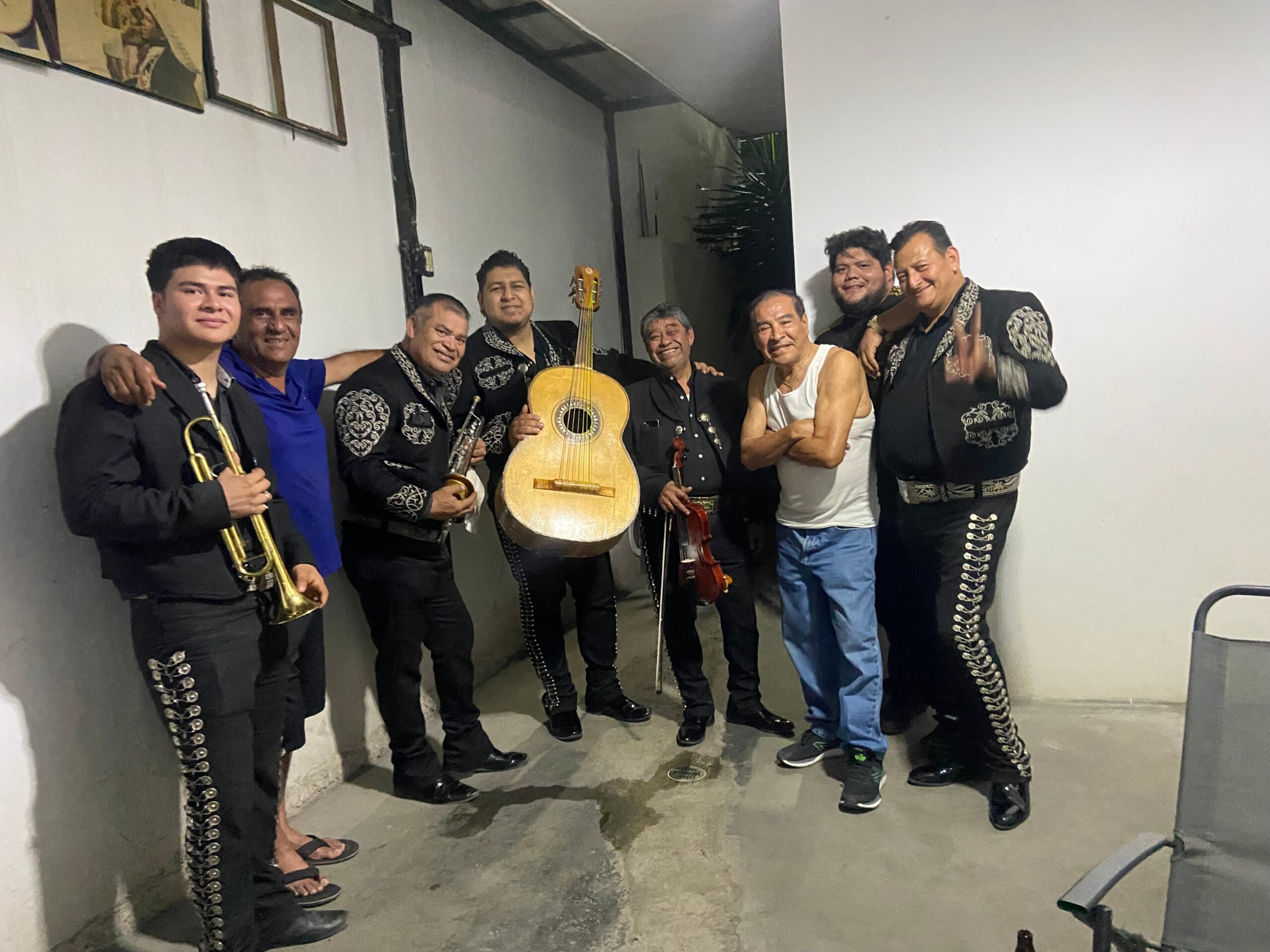 Group of mariachi musicians posing with instruments in a plain hallway, wearing black outfits with silver embroidery.