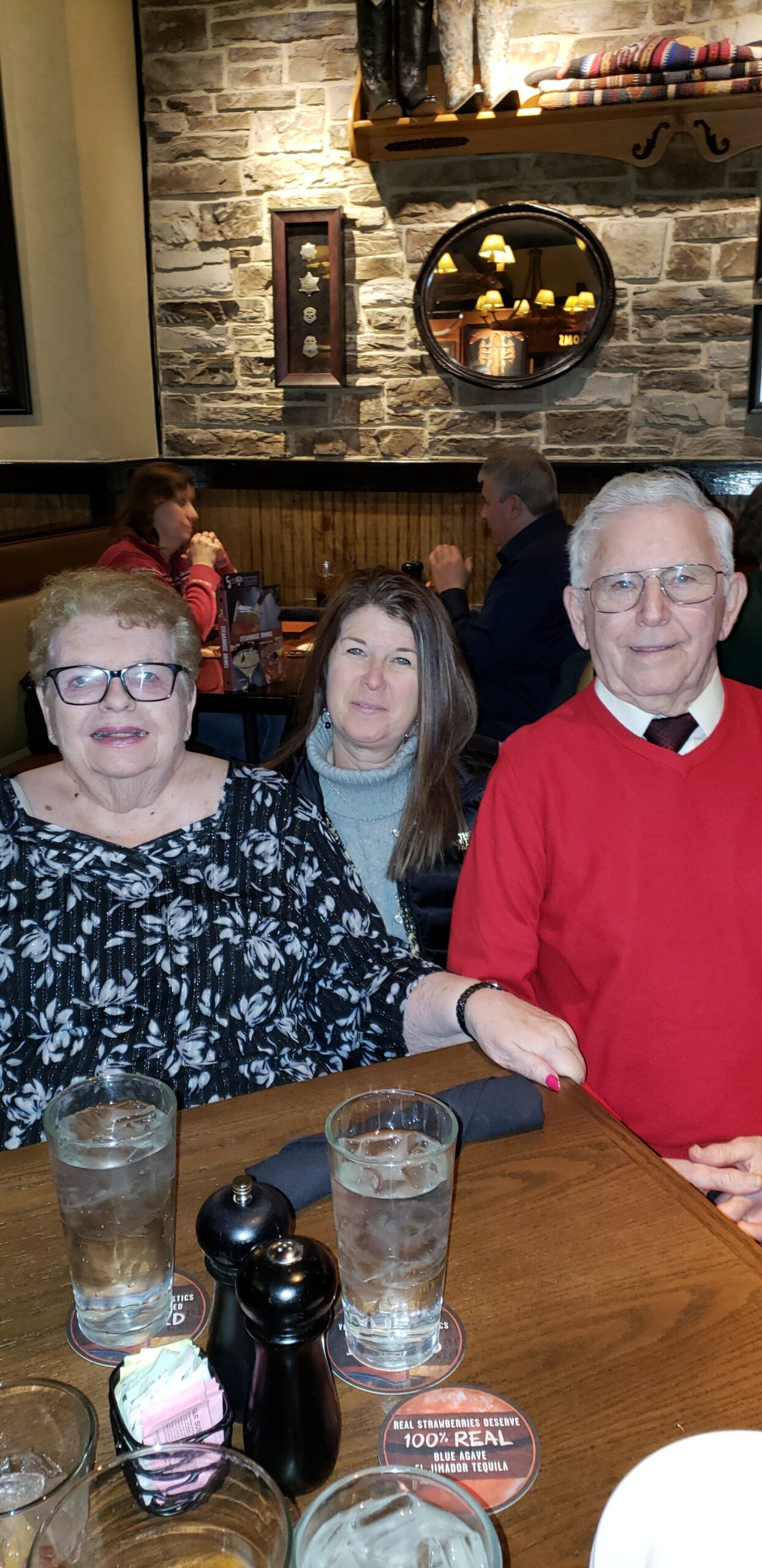 Three adults posing at a restaurant table: elderly woman with glasses on the left, middle-aged woman in the center, and an older man in a red sweater on the right, with drinks on the table in the foreground.
