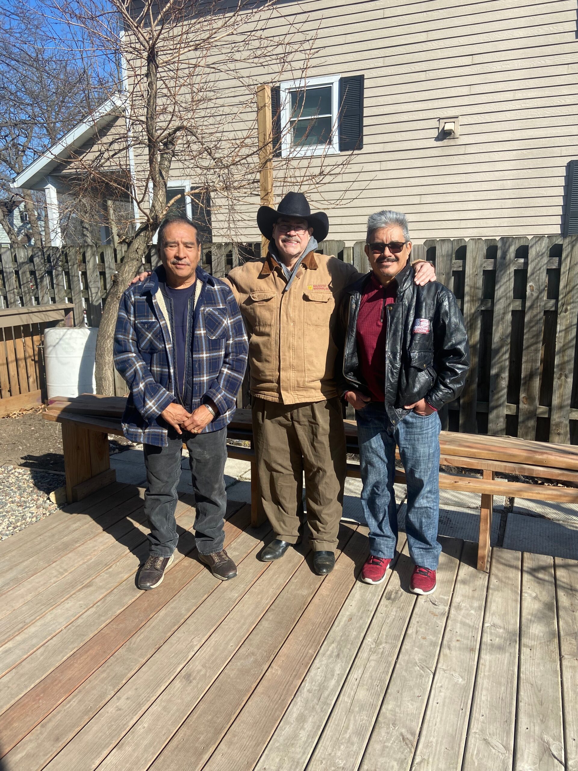 Three men stand together on a wooden deck in a backyard, posing for a photo.” (informative)