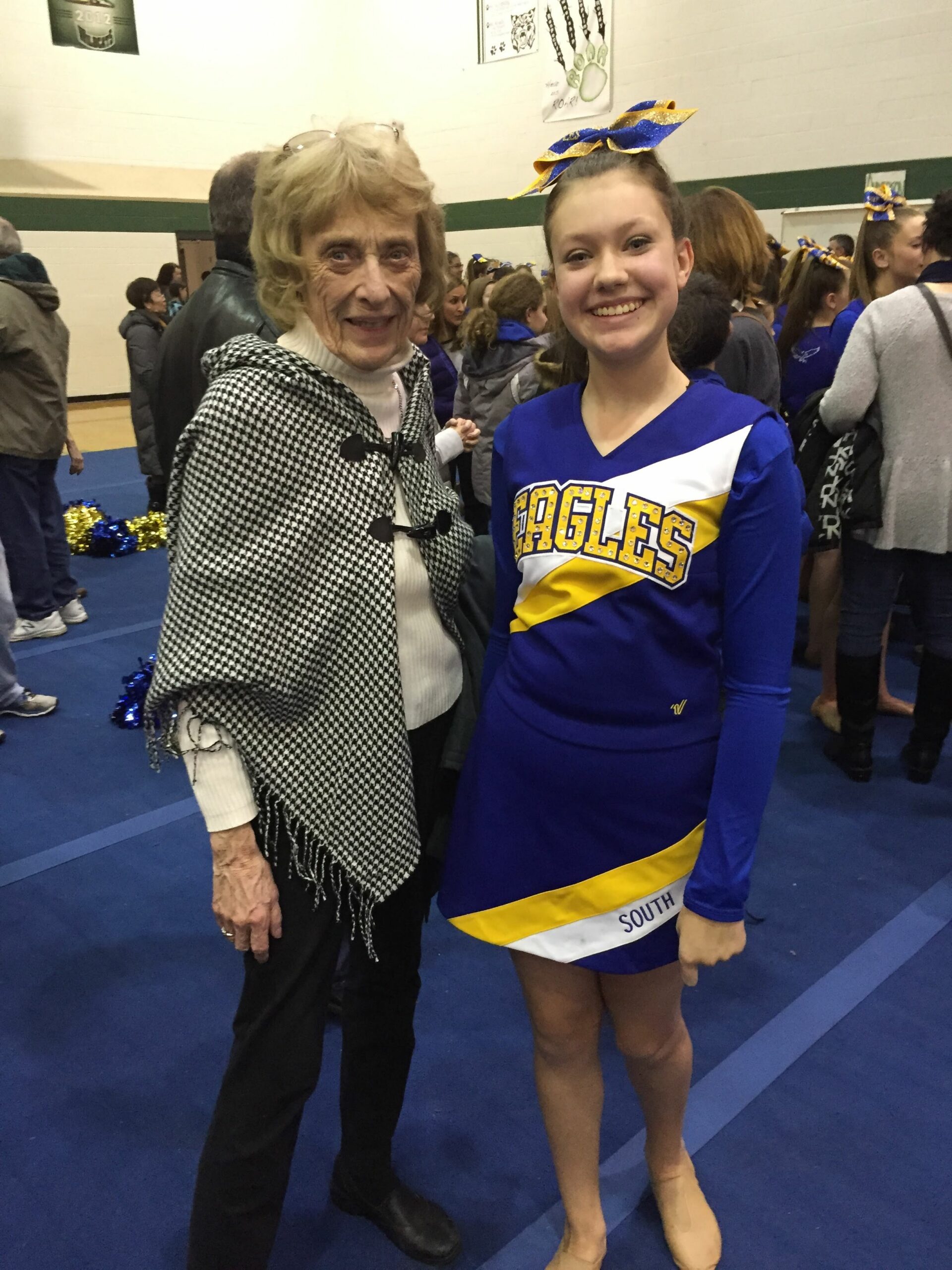 Smiling elderly woman in a black-and-white houndstooth shawl stands beside a teenage cheerleader in a blue and yellow Eagles uniform with a matching bow in a gym.