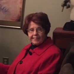 Portrait of an elderly woman with short brown hair and glasses, wearing a red coat, seated indoors and facing the camera.