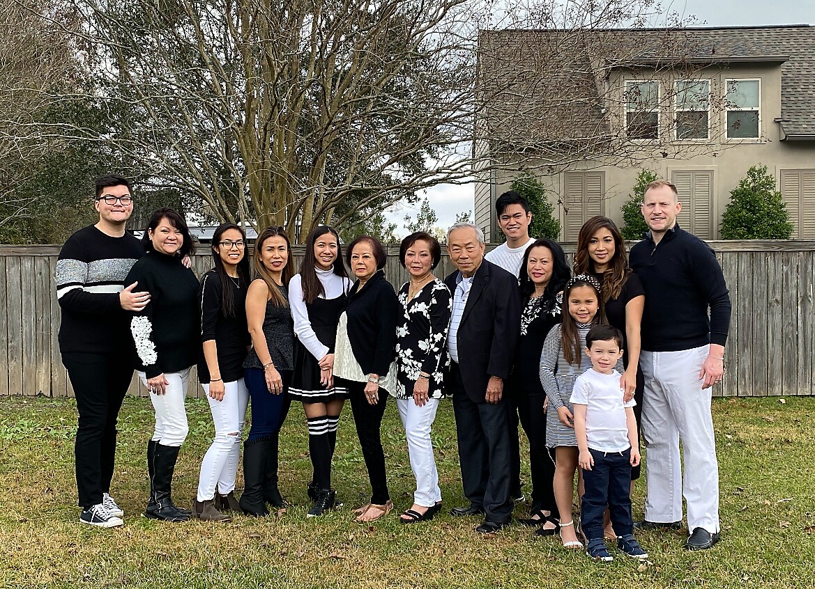 Large family group posing outdoors on a lawn, dressed in black and white, in front of a wooden fence and beige house.