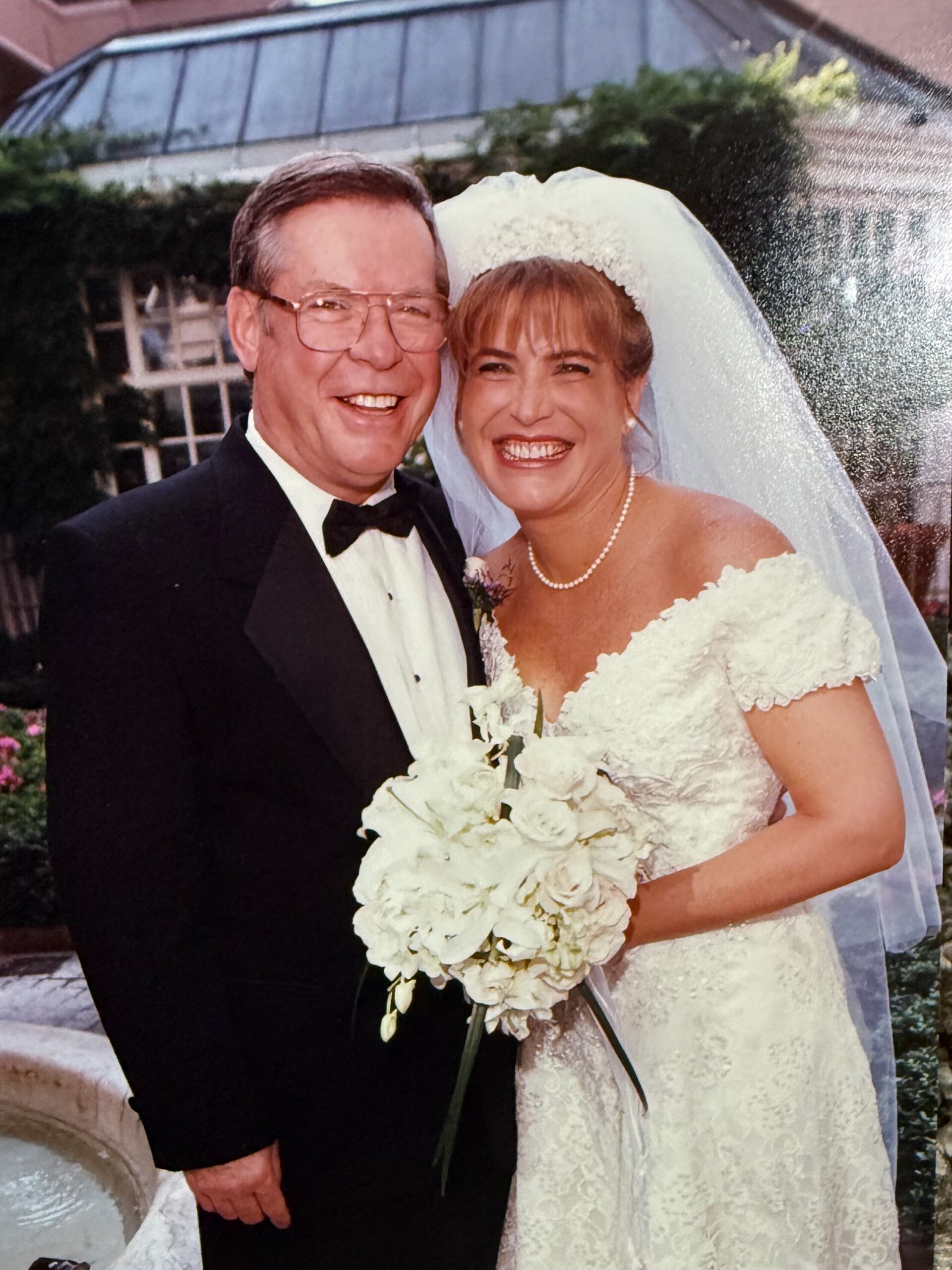 Smiling bride in lace wedding gown and veil beside groom in a tuxedo outdoors, holding a white bouquet of roses.