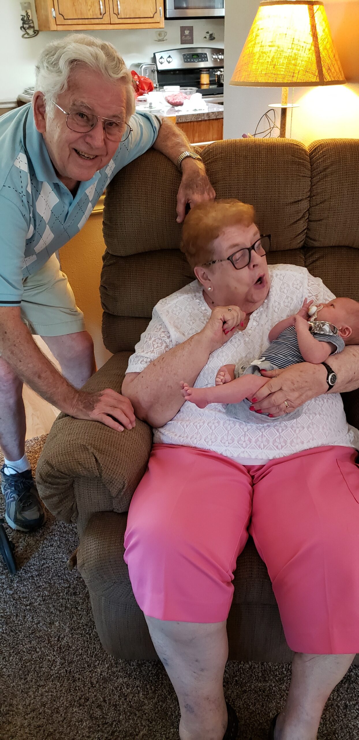 Two elderly adults crowding around a brown recliner: a smiling man rests his arm on the chair while an elderly woman in a white lace top holds a newborn baby wearing a striped outfit, in a cozy living room setting