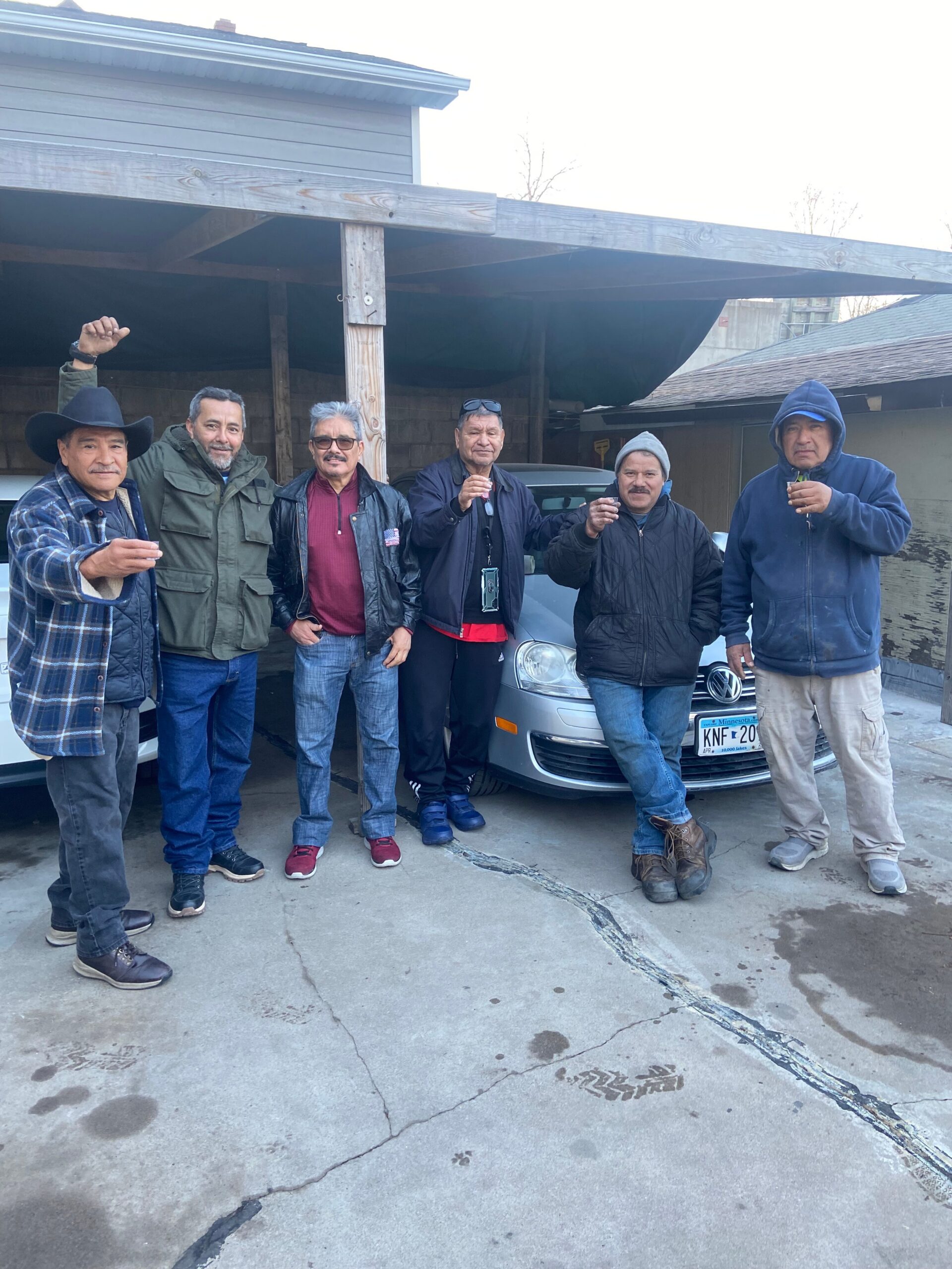Six men stand together under a wooden carport, holding drinks and smiling beside a silver car.