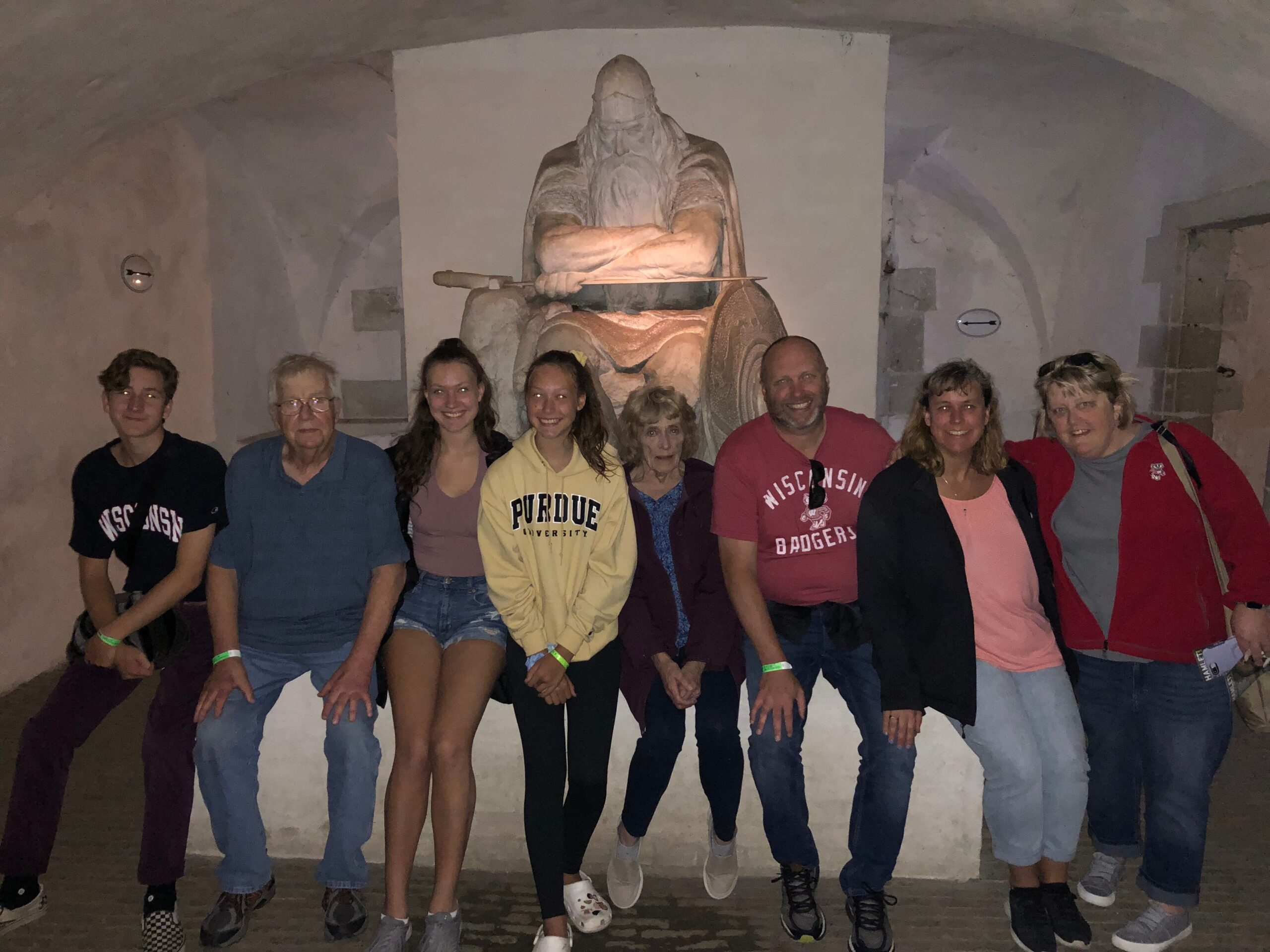 Group of nine people posing in a dim cave with a large stone statue behind them, some wearing university apparel and casual clothing, smiling for the photo.