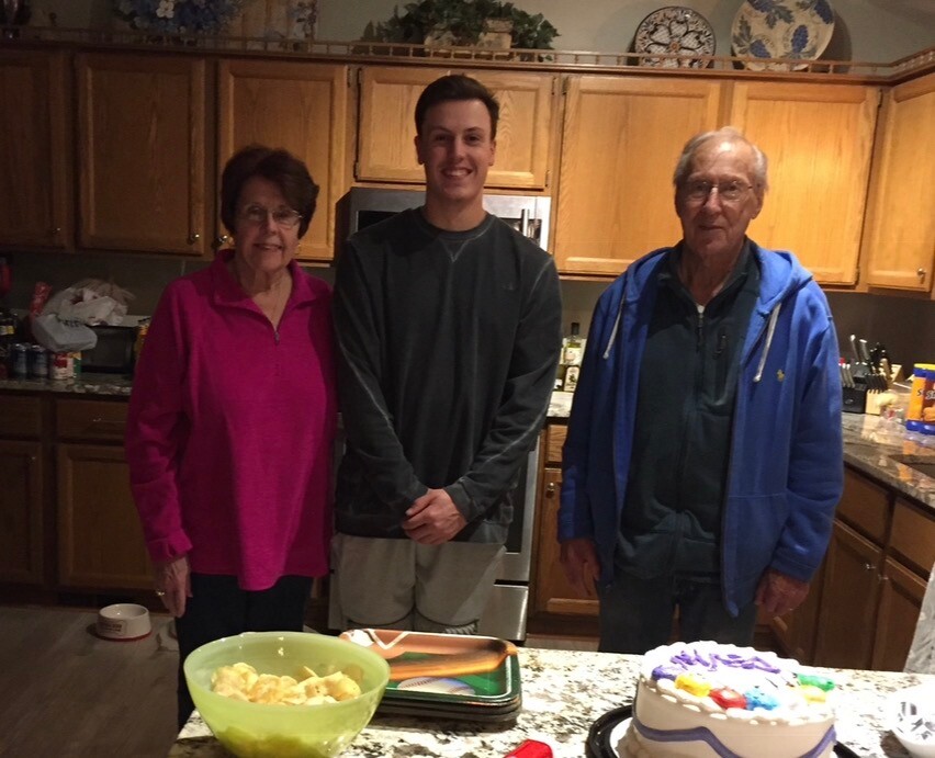 Three people stand in a kitchen beside a frosting cake on the counter, posing for a photo.