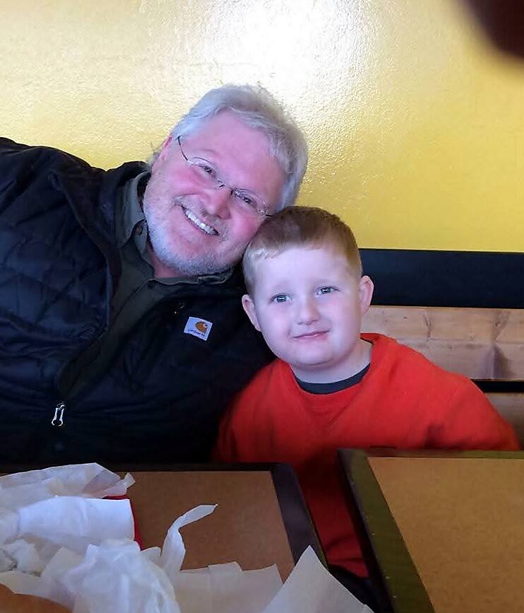 Smiling man with gray hair and glasses sits next to a young boy in a red shirt at a restaurant, yellow wall behind them.