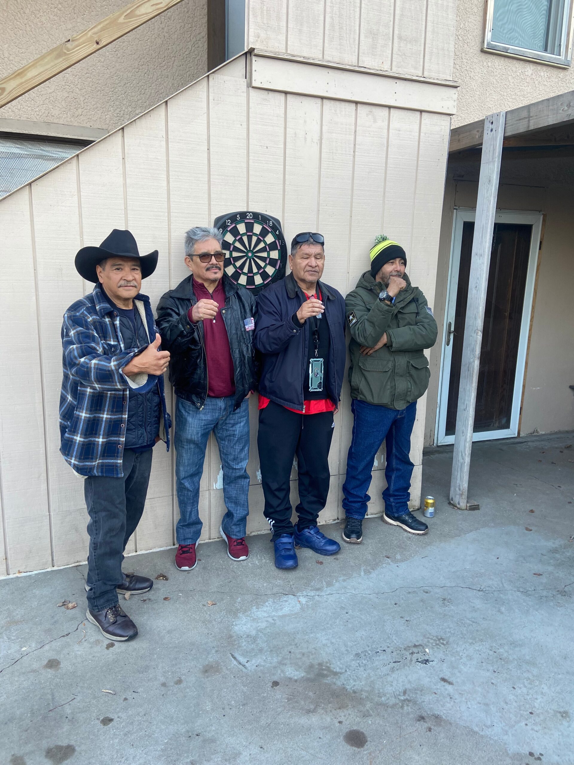 Four men pose outdoors beside a wall with a mounted dartboard; one wears a cowboy hat and they each hold drinks or gesture.