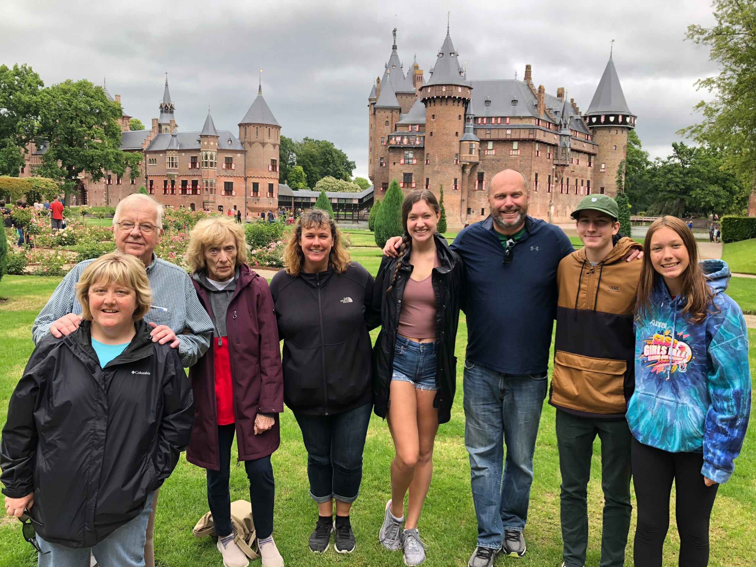 Group of smiling people posing on a grassy lawn in front of a large castle with towers and red-brick walls.