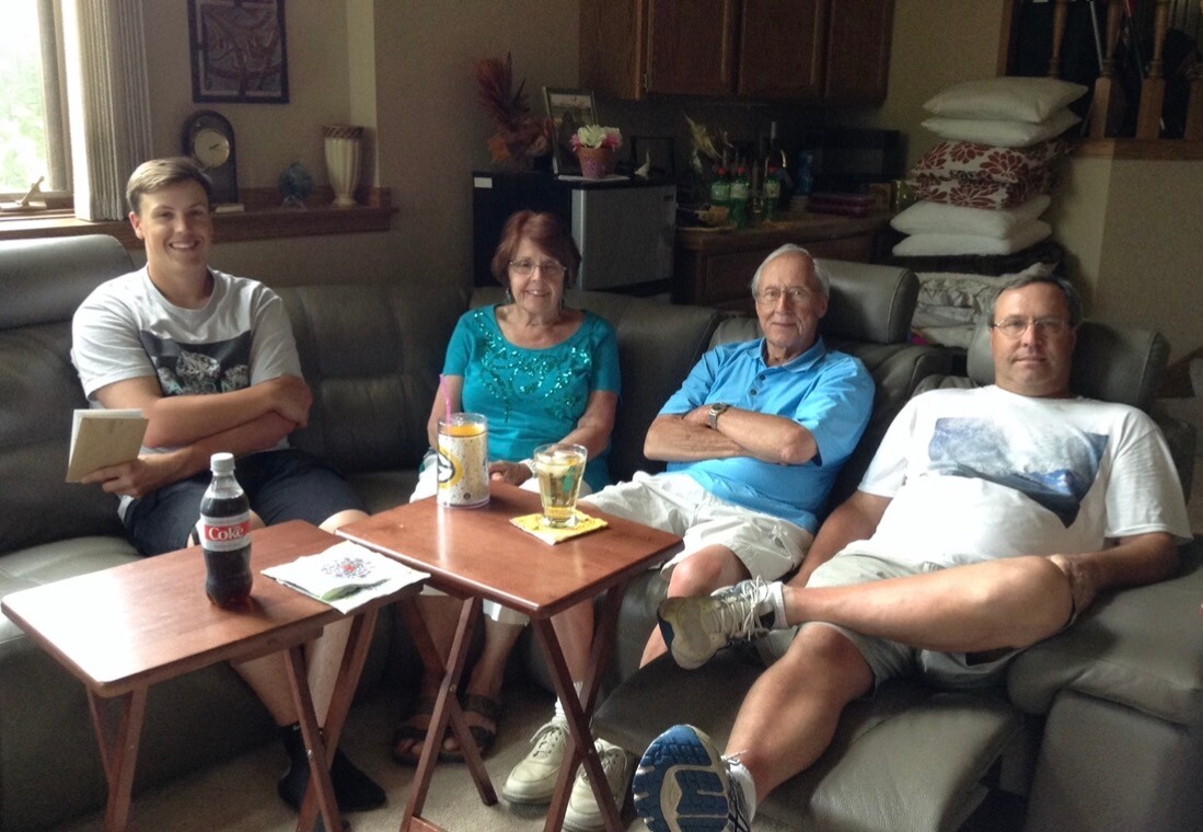Four people relax on a living room sofa with drinks on a small table: a young man on the left holds a notebook, others smile nearby.
