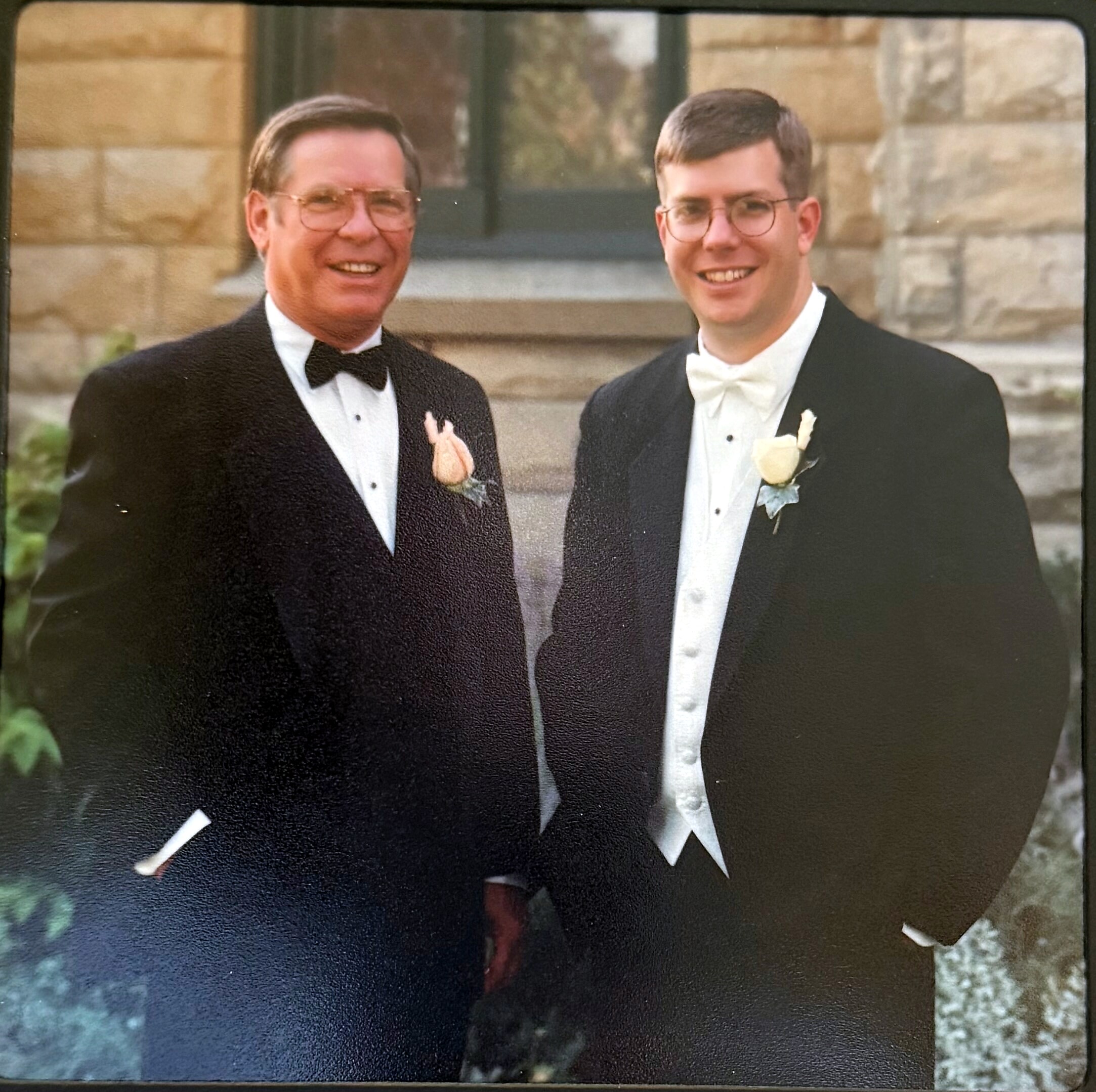 Two men in tuxedos posing together outdoors in front of a stone building, both smiling.