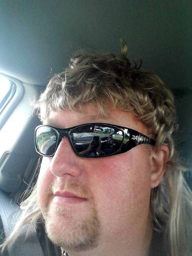Man with a mullet hairstyle wearing black sunglasses in a car, reflection of the road visible in the lenses.
