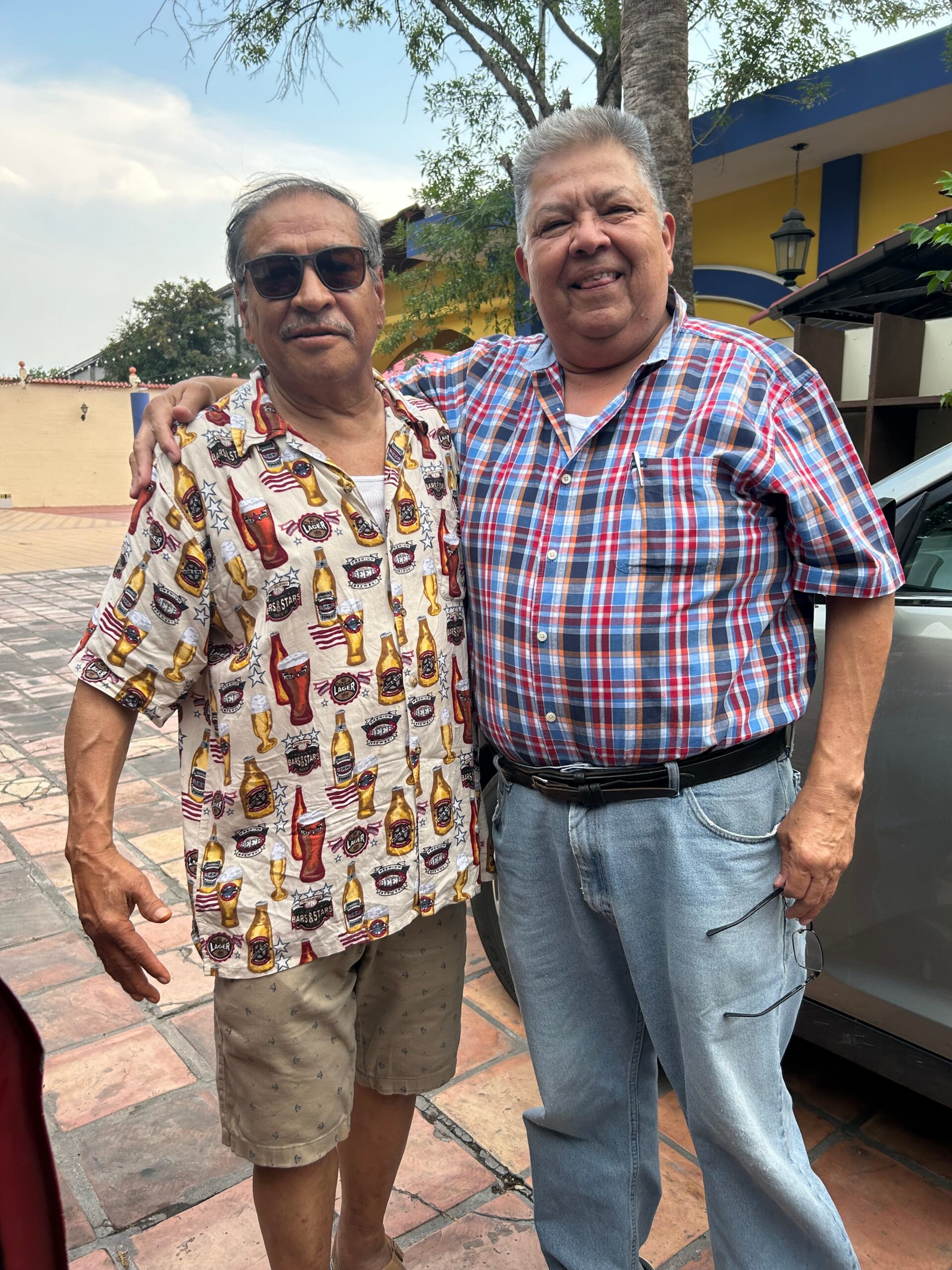 Two men stand together outdoors, one wearing sunglasses and a beer-themed shirt, the other in a blue plaid shirt, smiling with a colorful building behind them.