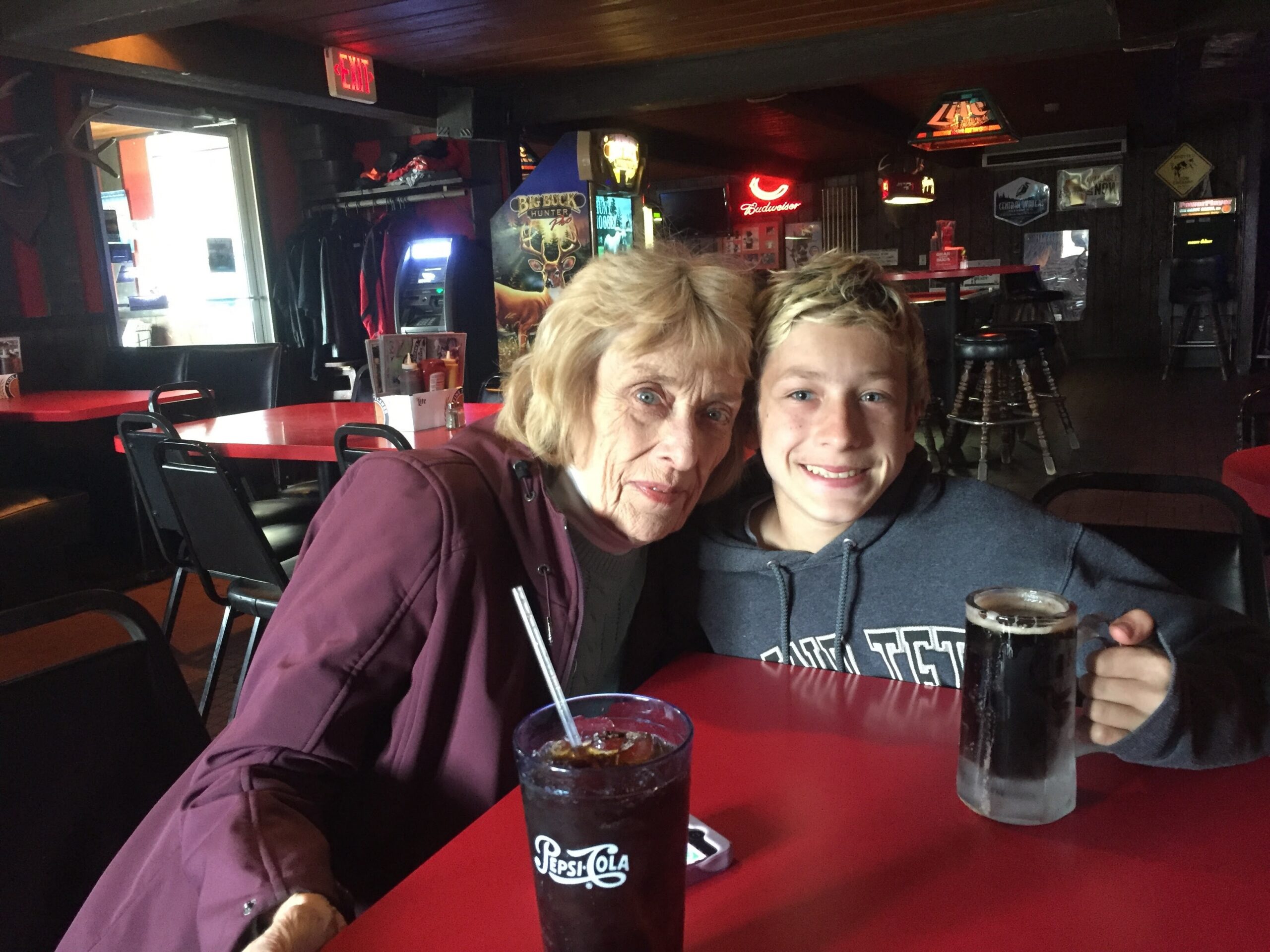 Smiling woman and boy posing for a selfie at a red-table, retro-style bar with drinks in the foreground and neon signs in the background.