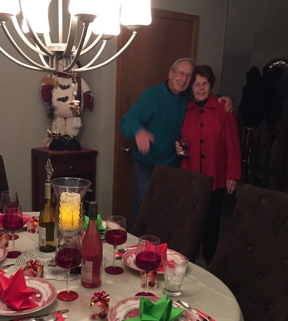 An elderly couple posing together by a festive dinner table, smiling, with wine glasses and Christmas decorations nearby.