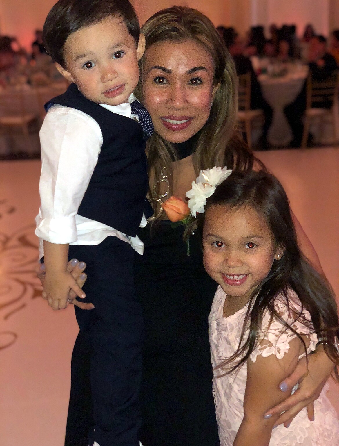 Smiling woman in a black dress with a young boy in a vest and a girl in a pink lace dress at a formal event.