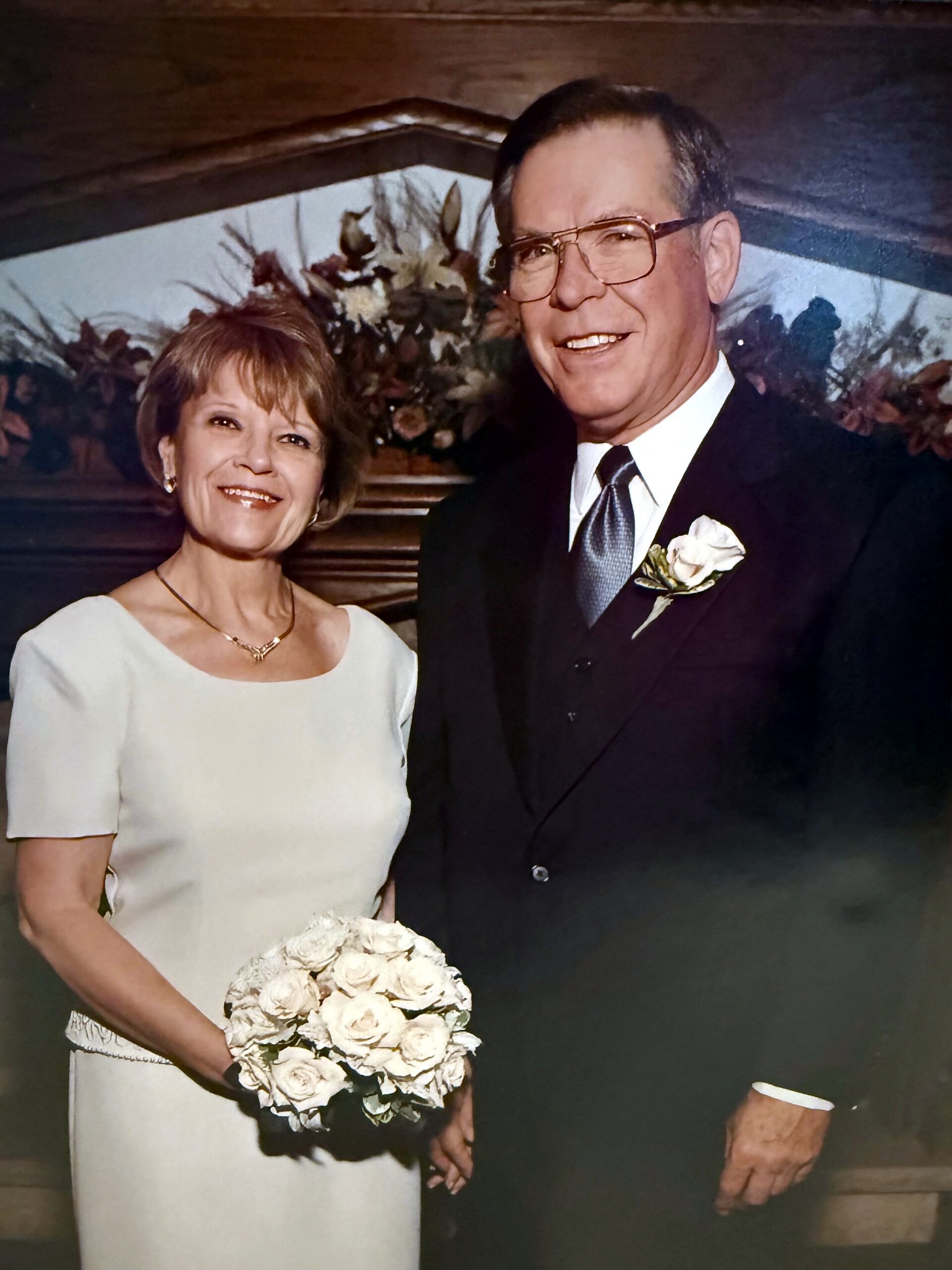 Smiling couple in formal wear at a wedding; the woman holds a bouquet of white roses.