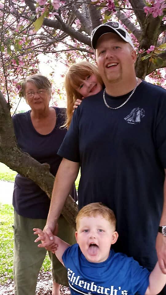 A family posing outdoors under pink blossoming trees: a man in a cap, an older woman, a girl behind him, and a toddler in front sticking out his tongue.