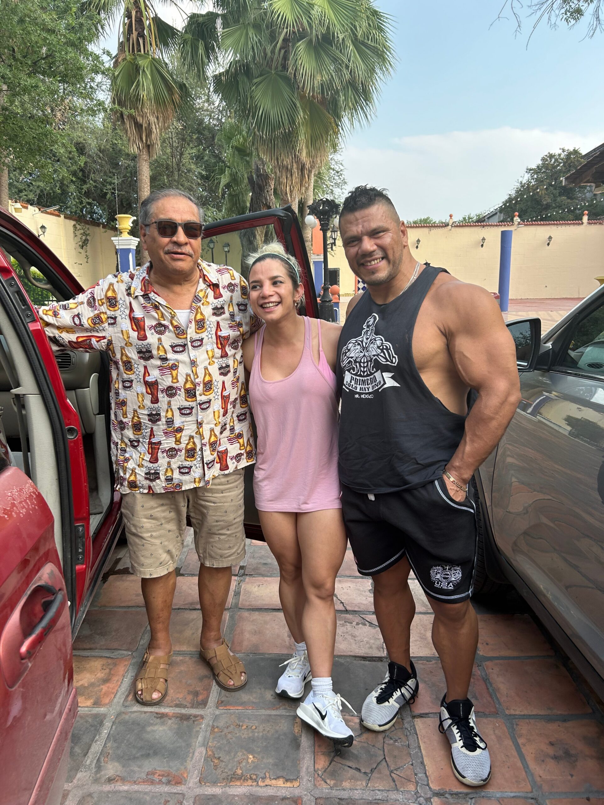 Three people pose together beside parked cars outdoors, smiling with palm trees and a beige wall in the background.