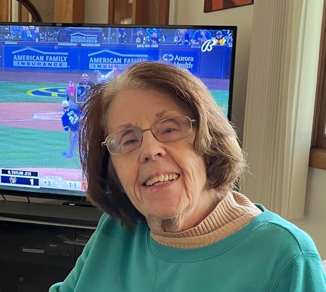 Smiling elderly woman with glasses sits in a living room, with a baseball game on the TV behind her.
