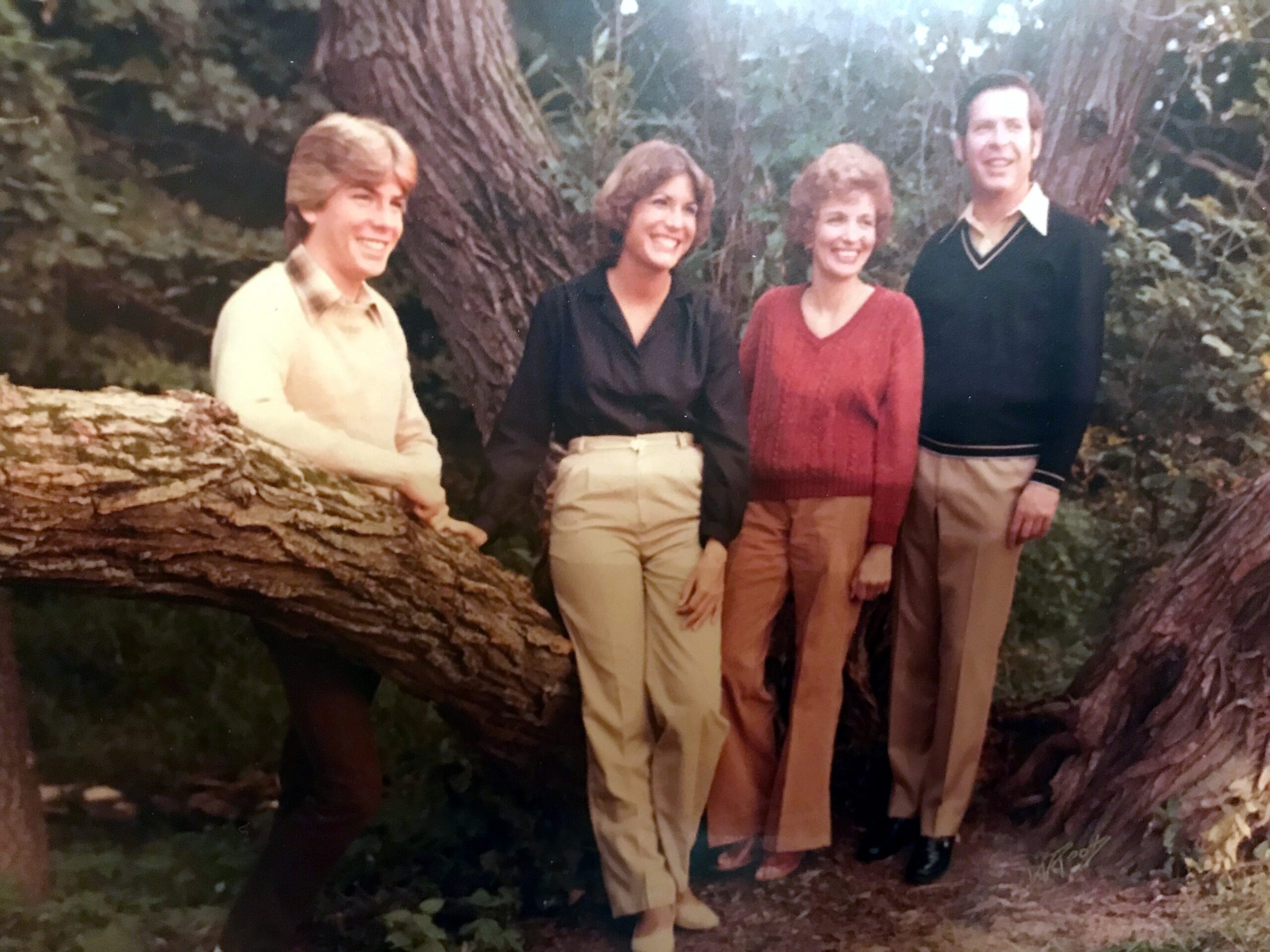 Four adults posing on a large fallen tree in a forest, smiling at the camera.